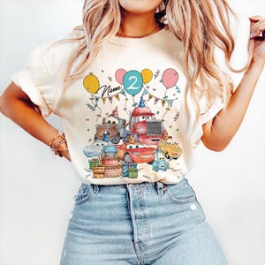 May include: Cream-colored t-shirt featuring a colorful graphic of animated cars, balloons, and a birthday cake. The number "2" is displayed on a balloon, with the word "Name" above. The design includes party hats and gift boxes.