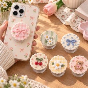 May include: A clear smartphone case with a pink floral and bow pattern, featuring a matching pink gingham bow phone grip. Five additional circular phone grips are displayed, featuring designs of daisies, a blue bow, cherries, a yellow gingham daisy, and a teddy bear.