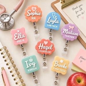 May include: A collection of eight colourful badge reels featuring various shapes and names including Sophie RN, Layla Lab Tech, Madison Teacher, Ella Pre-K, Hazel Counselor, Ivy Kinder, and Brooke X-Ray Tech, each with a clear plastic clip.