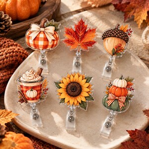 May include: Six autumn-themed badge reels featuring a plaid pumpkin with a bow, a vibrant orange maple leaf, an acorn, a coffee cup with whipped cream, a large yellow sunflower, and a stack of three pumpkins with a checkered ribbon.