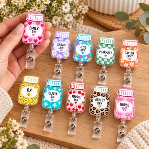 May include: A collection of ten acrylic badge reels shaped like medicine bottles, each featuring unique patterns including hearts, daisies, pills, checkers, smiley faces, crosses, cherries, leopard print, and bows, with personalized text labels.