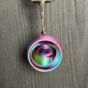 May include: A multi-colored, gradient-hued spherical fidget toy with concentric rings, suspended from a silver-toned metal lobster clasp attached to a black fabric strap. The object features a smooth, layered finish transitioning through shades of pink, blue, and yellow.