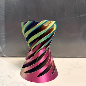 May include: A 3D printed vase featuring a spiraling, twisted geometric design. The object displays a vibrant, iridescent gradient finish that transitions through shades of magenta, purple, blue, green, and gold, creating a metallic sheen.