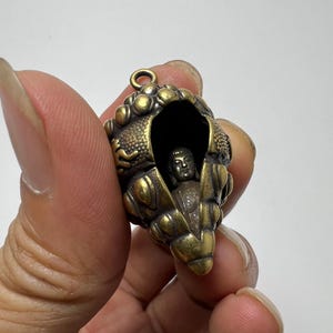 Peut inclure: Un pendentif en laiton en forme de coquillage texturé avec une petite figurine de Bouddha détaillée nichée à l'intérieur de l'ouverture. L'extérieur présente une finition dorée métallique avec des accents de patine sombre et une boucle.