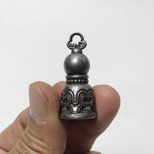 May include: A small, metallic, bell-shaped pendant with a weathered, dark gray finish. The object features a rounded top section, a central band of small raised spheres, and a flared base decorated with intricate, symmetrical, swirling embossed patterns.