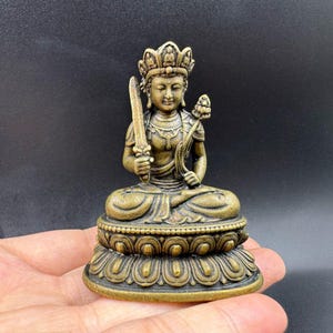 May include: A bronze-colored statue of a seated deity, adorned with a crown and intricate details. The figure holds a sword and a lotus flower, resting on a lotus-shaped base. The statue is approximately 3 inches tall, showcasing a serene expression.