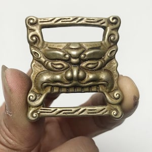 May include: A detailed, antique brass-colored buckle with a carved face design. The buckle features a rectangular shape with an open center and ornate scrollwork around the edges. The face has prominent eyes, a nose, and a mouth.