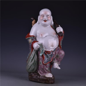 May include: A porcelain figurine of a smiling, bald figure with a large belly, wearing patterned robes in shades of pink, green, and blue. The figure carries a sack over one shoulder and a staff in the other hand, standing on a brown base.