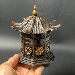 May include: A dark brown metal incense burner shaped like a traditional pagoda with a tiered roof, a finial top, and openwork lattice walls featuring circular gold-coloured floral medallions and geometric diamond-shaped accents on a hexagonal base.