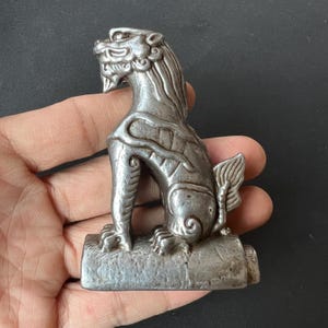May include: A silver-toned metal figurine of a mythical creature, likely a lion or dragon, depicted in a seated position on a rectangular base. The sculpture features intricate embossed details, a flowing mane, and a curled tail, with a weathered finish.