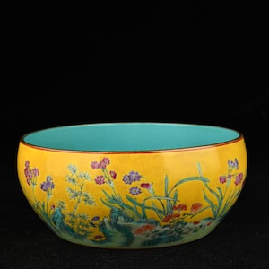 Pode incluir: Uma tigela de cerâmica redonda com um exterior amarelo vibrante apresentando um design floral pintado à mão com elementos botânicos roxos, vermelhos e verdes e formações rochosas. O interior da tigela é de uma cor azul turquesa lisa.