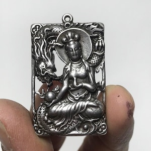 May include: A silver-colored rectangular pendant featuring a seated figure with a halo, holding a sphere, and a dragon on the left side. The pendant has a loop at the top for a chain. The design is intricate and detailed.