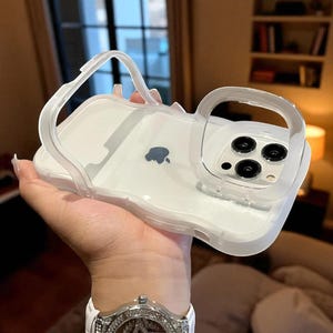 May include: Transparent phone case with a handle. The case is white with a clear handle and a clear border around the camera. The Apple logo is visible on the back of the case.