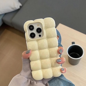 Puede incluir: Una funda de teléfono de color crema con un diseño acolchado y abultado. La funda tiene forma cuadrada con bordes redondeados y un recorte para la cámara. La funda está sujeta por una persona. Una pequeña taza gris está sobre una superficie clara.