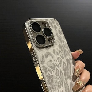 May include: A close-up of a smartphone case with a gray leopard print design. The case has a gold-colored frame and a silver camera lens protector. The case is designed to protect the phone.