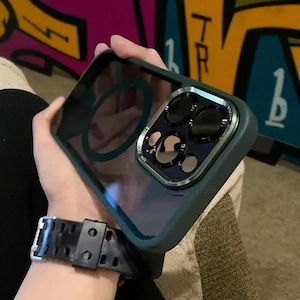 Puede incluir: Una funda transparente para teléfono con borde verde oscuro y protector de lente de cámara plateado. La funda tiene un anillo magnético circular en la parte posterior. La palabra "STUBIA" está impresa en la parte inferior.