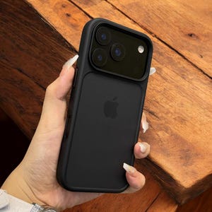May include: Black phone case with a matte finish, featuring a triple-lens camera cutout and the Apple logo. The case is designed to fit a smartphone and offers protection. The case is held against a wooden surface.