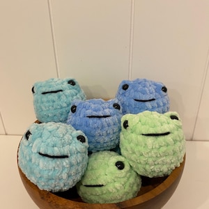 May include: A wooden bowl filled with seven plush frog toys in shades of blue and green. Each frog has black eyes and a stitched black smile. The frogs are made of a soft, textured material.