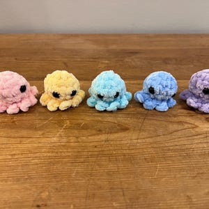 May include: Five small, plush octopuses in a row, each a different pastel color: pink, yellow, light blue, medium blue, and purple. They have black eyes and are sitting on a wooden surface.