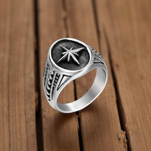 May include: A silver-colored ring with a starburst design set in a dark gray oval. The ring has a detailed pattern on the sides and a polished finish. The ring is displayed on a wooden surface.