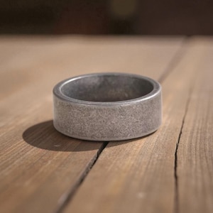 May include: A close-up of a thick, silver-colored ring with a textured surface. The ring is cylindrical and sits on a wooden surface. The ring's color is a muted gray, and the texture gives it a rustic appearance. The ring is a jewelry item.