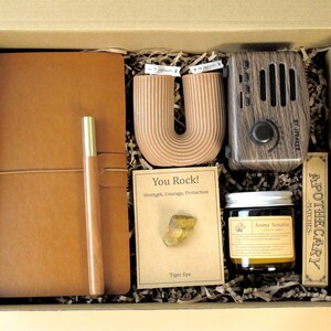 Peut inclure: Coffret cadeau avec un journal en cuir, un stylo, une bougie en forme de U, un haut-parleur en bois, une bougie en pot, des allumettes et une carte "You Rock!" avec une pierre oeil de tigre.
