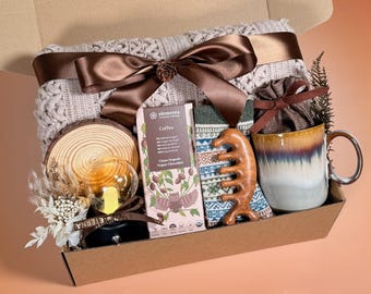 May include: A gift box containing a knitted blanket, coffee, socks, a wooden comb, and a mug. The box is tied with a brown satin ribbon and a pine cone. The mug has a brown handle and a blue and brown glaze.