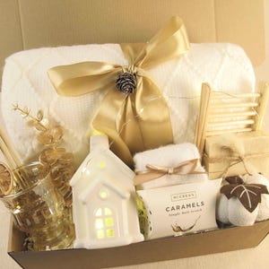 Puede incluir: Una cesta de regalo llena de una manta blanca atada con una cinta dorada, un candelabro blanco con forma de casa, un vaso con una rodaja de naranja seca y otros art&iacute;culos. La cesta incluye una peque&ntilde;a tabla de lavar de madera y una caja de caramelos.