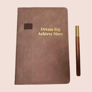 Puede incluir: Un diario marrón con el texto dorado "Dream Big Achieve More" y un bolígrafo a juego. El diario tiene un borde cosido y una banda elástica. El bolígrafo tiene una parte superior dorada y un cuerpo de madera marrón.