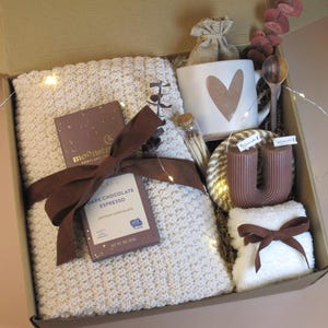 Puede incluir: Una caja de regalo llena de una manta de punto color crema, una taza con un dise&ntilde;o de coraz&oacute;n, chocolate negro, una vela y otros art&iacute;culos. La caja est&aacute; atada con una cinta marr&oacute;n e incluye luces de hadas para una est&eacute;tica acogedora.