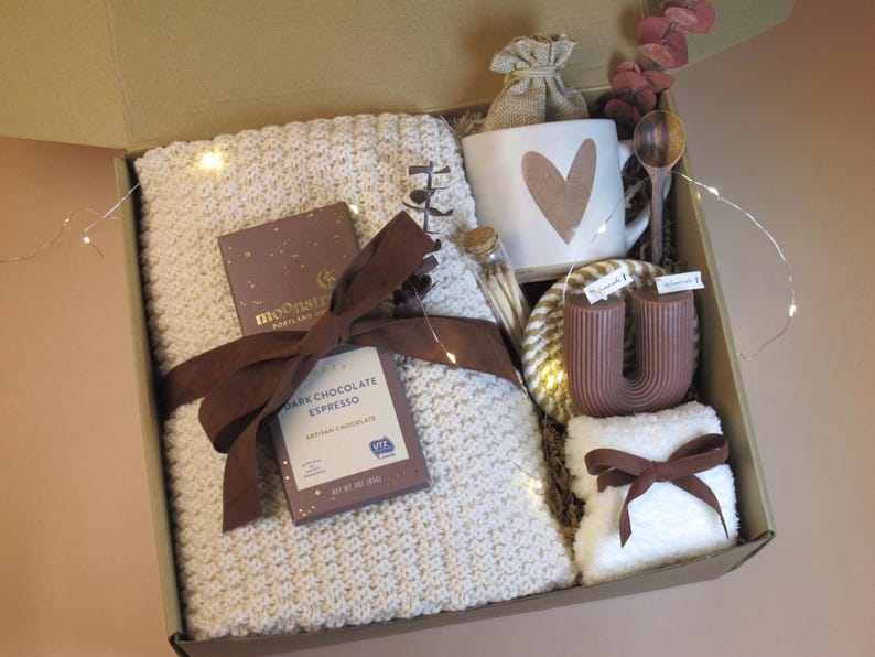 Puede incluir: Una caja de regalo llena de una manta de punto color crema, una taza con un dise&ntilde;o de coraz&oacute;n, chocolate negro, una vela y otros art&iacute;culos. La caja est&aacute; atada con una cinta marr&oacute;n e incluye luces de hadas para una est&eacute;tica acogedora.
