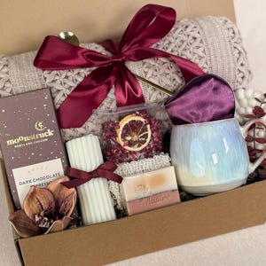 Puede incluir: Una caja de regalo llena de una manta de punto beige, un lazo burdeos, una taza con un esmalte azul y blanco, chocolate negro, una vela, jab&oacute;n y flores secas. La caja es marr&oacute;n y los art&iacute;culos est&aacute;n dentro.