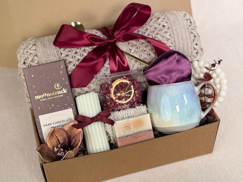 Puede incluir: Una caja de regalo llena de una manta de punto beige, un lazo burdeos, una taza con un esmalte azul y blanco, chocolate negro, una vela, jab&oacute;n y flores secas. La caja es marr&oacute;n y los art&iacute;culos est&aacute;n dentro.