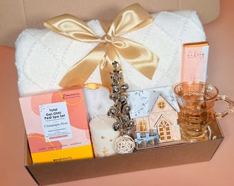 May include: A gift box containing a white blanket, gold ribbon, spa set, candle, decorative house, glass mug, and chocolate bar. The brown box sits on a peach surface. The spa set is for a Champagne Hour.
