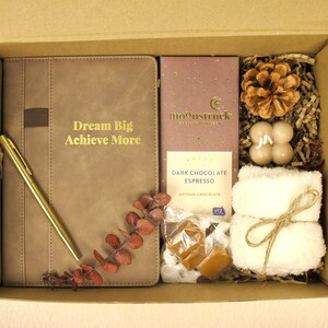 Puede incluir: Caja de regalo llena de un diario marr&oacute;n con el texto "Dream Big Achieve More", un bol&iacute;grafo dorado, una tableta de chocolate negro, una pi&ntilde;a, velas, una peque&ntilde;a toalla blanca y caramelos.