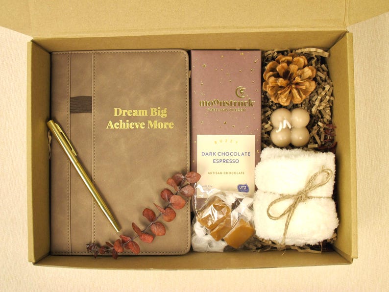 Puede incluir: Caja de regalo llena de un diario marr&oacute;n con el texto "Dream Big Achieve More", un bol&iacute;grafo dorado, una tableta de chocolate negro, una pi&ntilde;a, velas, una peque&ntilde;a toalla blanca y caramelos.