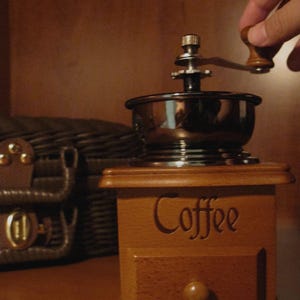 Peut inclure: Un moulin &agrave; caf&eacute; en bois avec un tiroir et le mot "Coffee" grav&eacute; sur le devant. Le moulin a un dessus en m&eacute;tal et une manivelle en bois. Une main tourne la manivelle. Une bo&icirc;te en cuir marron est &agrave; l'arri&egrave;re-plan.