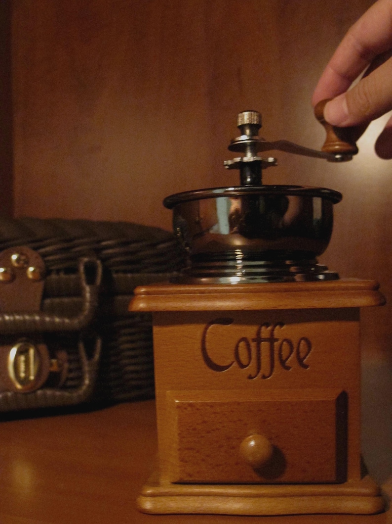 Peut inclure: Un moulin &agrave; caf&eacute; en bois avec un tiroir et le mot "Coffee" grav&eacute; sur le devant. Le moulin a un dessus en m&eacute;tal et une manivelle en bois. Une main tourne la manivelle. Une bo&icirc;te en cuir marron est &agrave; l'arri&egrave;re-plan.