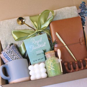 Puede incluir: Una caja de regalo llena de art&iacute;culos de cuidado personal. Incluye una taza azul, un diario de cuero marr&oacute;n, un bol&iacute;grafo, una toalla peque&ntilde;a, t&eacute; de hierbas, una vela y una cuchara decorativa. La caja est&aacute; atada con una cinta verde.