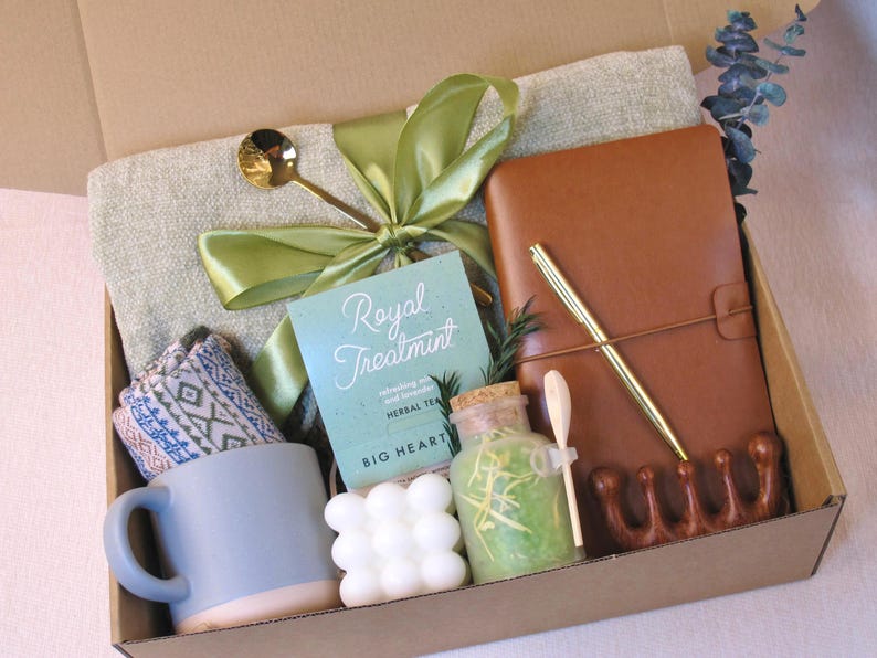 Puede incluir: Una caja de regalo llena de art&iacute;culos de cuidado personal. Incluye una taza azul, un diario de cuero marr&oacute;n, un bol&iacute;grafo, una toalla peque&ntilde;a, t&eacute; de hierbas, una vela y una cuchara decorativa. La caja est&aacute; atada con una cinta verde.