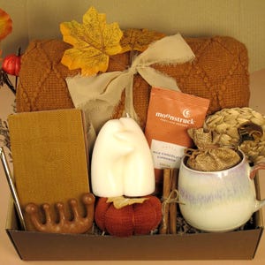 Puede incluir: Una cesta de regalo con tem&aacute;tica oto&ntilde;al con una manta marr&oacute;n, una vela, una taza, un diario y un paquete de chocolate caliente Moonstruck. La cesta est&aacute; decorada con hojas de oto&ntilde;o y un lazo.