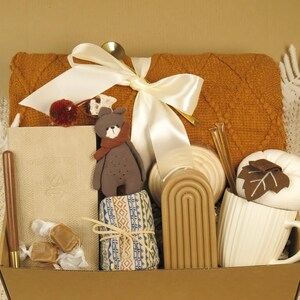 Puede incluir: Una caja de regalo llena de art&iacute;culos acogedores. Incluye una manta naranja texturizada, un oso de peluche, un cuaderno, una taza con una hoja, velas y caramelos envueltos. Una cinta blanca y una cuchara dorada a&ntilde;aden un toque decorativo.