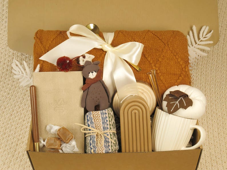Puede incluir: Una caja de regalo llena de art&iacute;culos acogedores. Incluye una manta naranja texturizada, un oso de peluche, un cuaderno, una taza con una hoja, velas y caramelos envueltos. Una cinta blanca y una cuchara dorada a&ntilde;aden un toque decorativo.