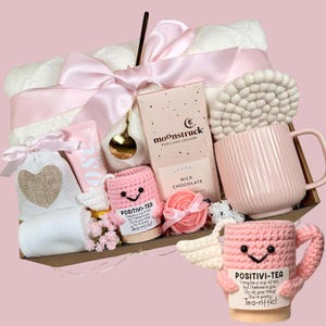Pode incluir: Uma cesta de presente com uma caneca rosa, chocolate, uma pequena bolsa, loção, uma capa de caneca de crochê, uma toalha branca e um laço rosa. A capa da caneca tem o texto "POSITIVI-TEA".