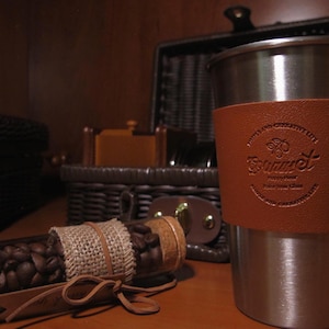 Peut inclure: Un mug de voyage en acier inoxydable avec un manchon en cuir marron, un tube en verre rempli de grains de caf&eacute; et un panier en osier. Le manchon en cuir porte le mot "Gourmet" en relief.