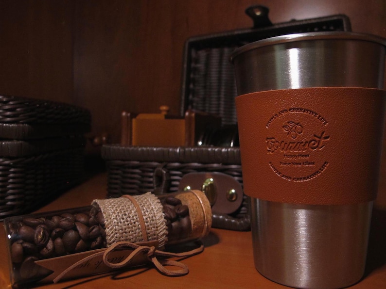 Peut inclure: Un mug de voyage en acier inoxydable avec un manchon en cuir marron, un tube en verre rempli de grains de caf&eacute; et un panier en osier. Le manchon en cuir porte le mot "Gourmet" en relief.
