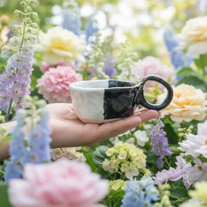 Puede incluir: Una pequeña taza de cerámica hecha a mano con un diseño bicolor que presenta un acabado blanco brillante en un lado y un acabado negro brillante en el otro. La taza tiene una forma orgánica irregular y un asa circular unida al lado negro.