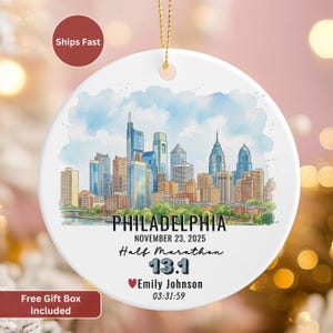 May include: White ceramic ornament featuring a watercolor illustration of the Philadelphia skyline. The ornament includes text that reads "PHILADELPHIA", "Half Marathon", "13.1", and the name "Emily Johnson". A gold string is attached for hanging.