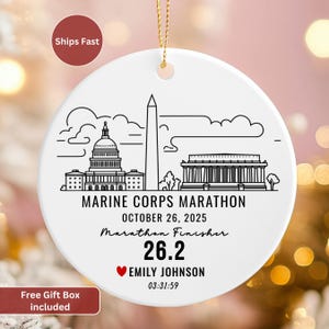 May include: White ceramic ornament with a gold string, featuring a black line drawing of Washington D.C. landmarks. Text includes "Marine Corps Marathon," date, distance, name, and time. A red heart is included.