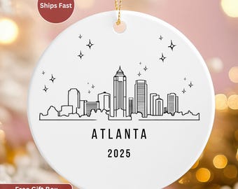 May include: A white, round ceramic ornament featuring a black line drawing of the Atlanta skyline. The word "ATLANTA" is printed below the skyline, with "2025" beneath it. The ornament has a gold string for hanging and star accents.